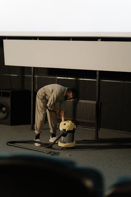 A woman using a yellow vacuum cleaner to clean a patterned area rug in a living room with wooden flooring and a red leather sofa, demonstrating surface cleaning and deep cleaning processes. The room i